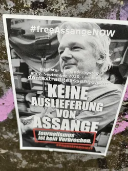 Black and white photo of Julian Assange with text overlay advocating for his release.  The sticker includes a website and date of a court hearing.