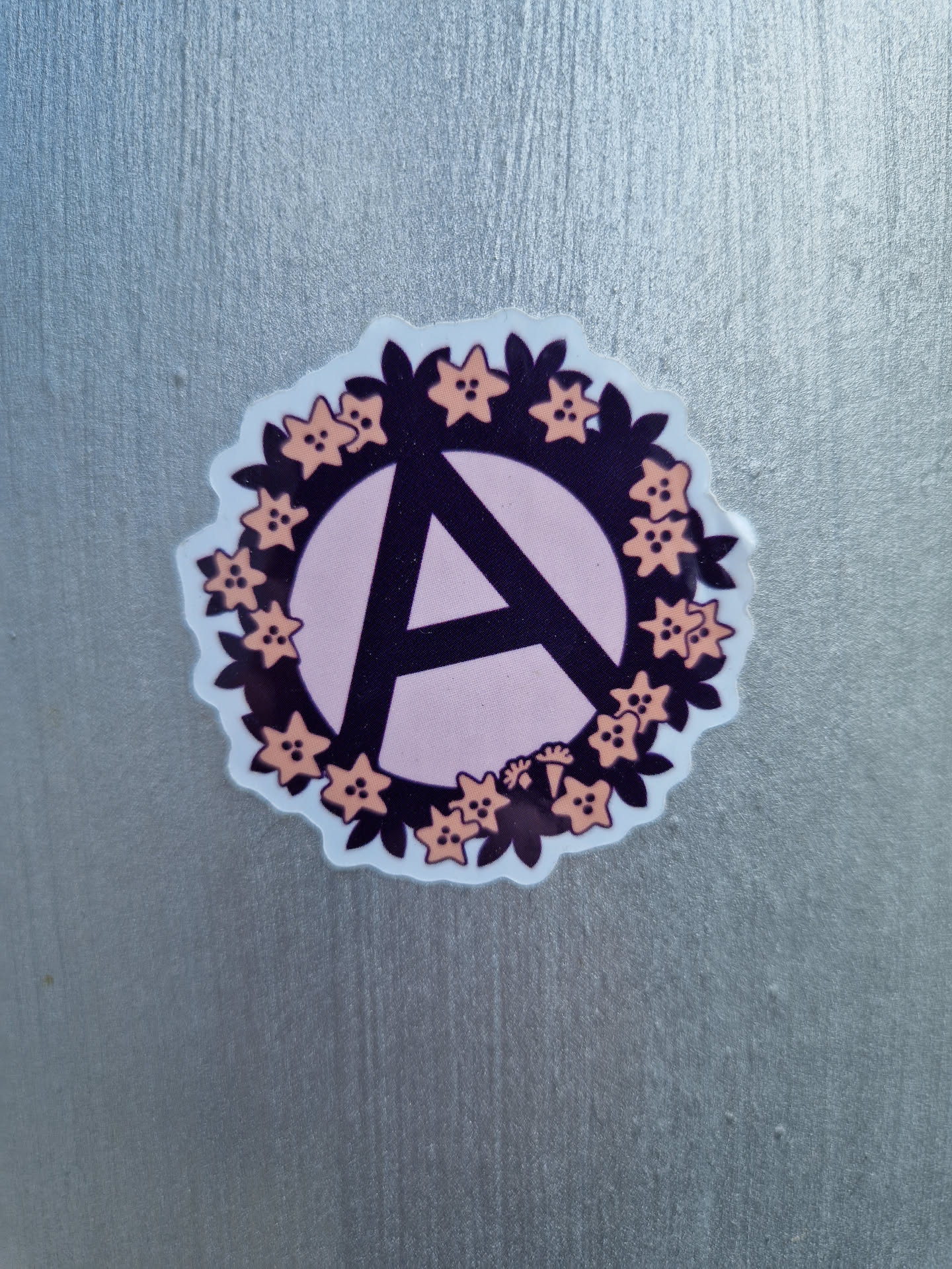 Round sticker with a pink anarchy symbol in the center, surrounded by a wreath of peach-colored flowers and dark purple leaves. The sticker is on a silver surface.