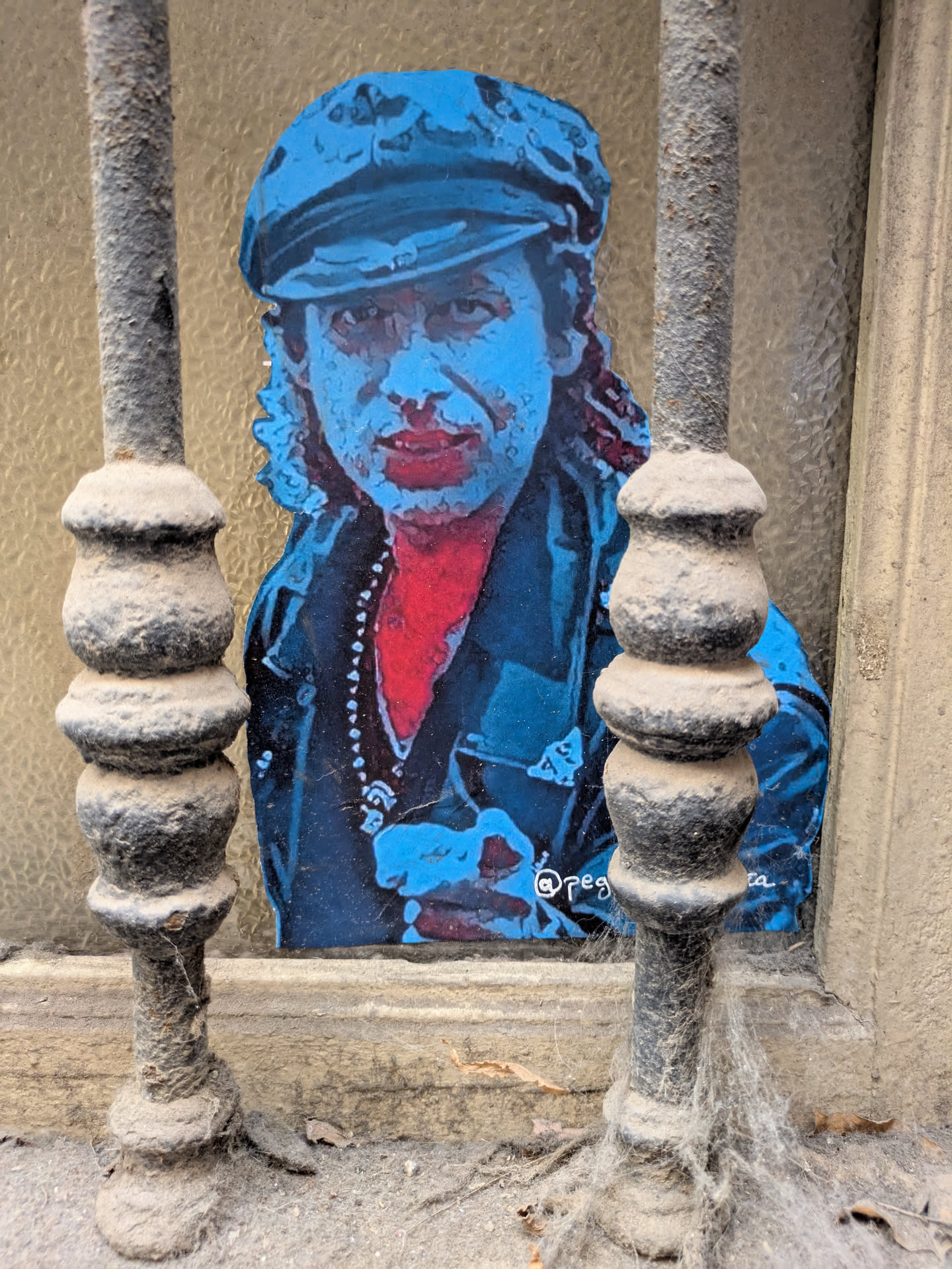 A sticker of a blue-skinned man with red lips and a red shirt, wearing a cap, is affixed to a window behind metal bars. The artwork, reminiscent of Mick Jagger, is detailed and appears somewhat faded. An Instagram handle is present at the bottom of the sticker.