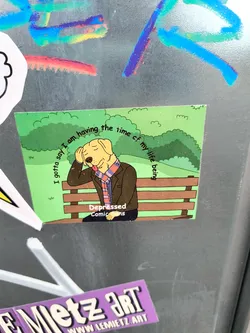 A sticker featuring a cartoon dog sitting on a park bench with text that says, "I gotta say I am having the time of my life being depressed." The sticker has a green background and is created by Depressed Comic Sans. There is also another sticker from www.lemietz.art visible in the photo.