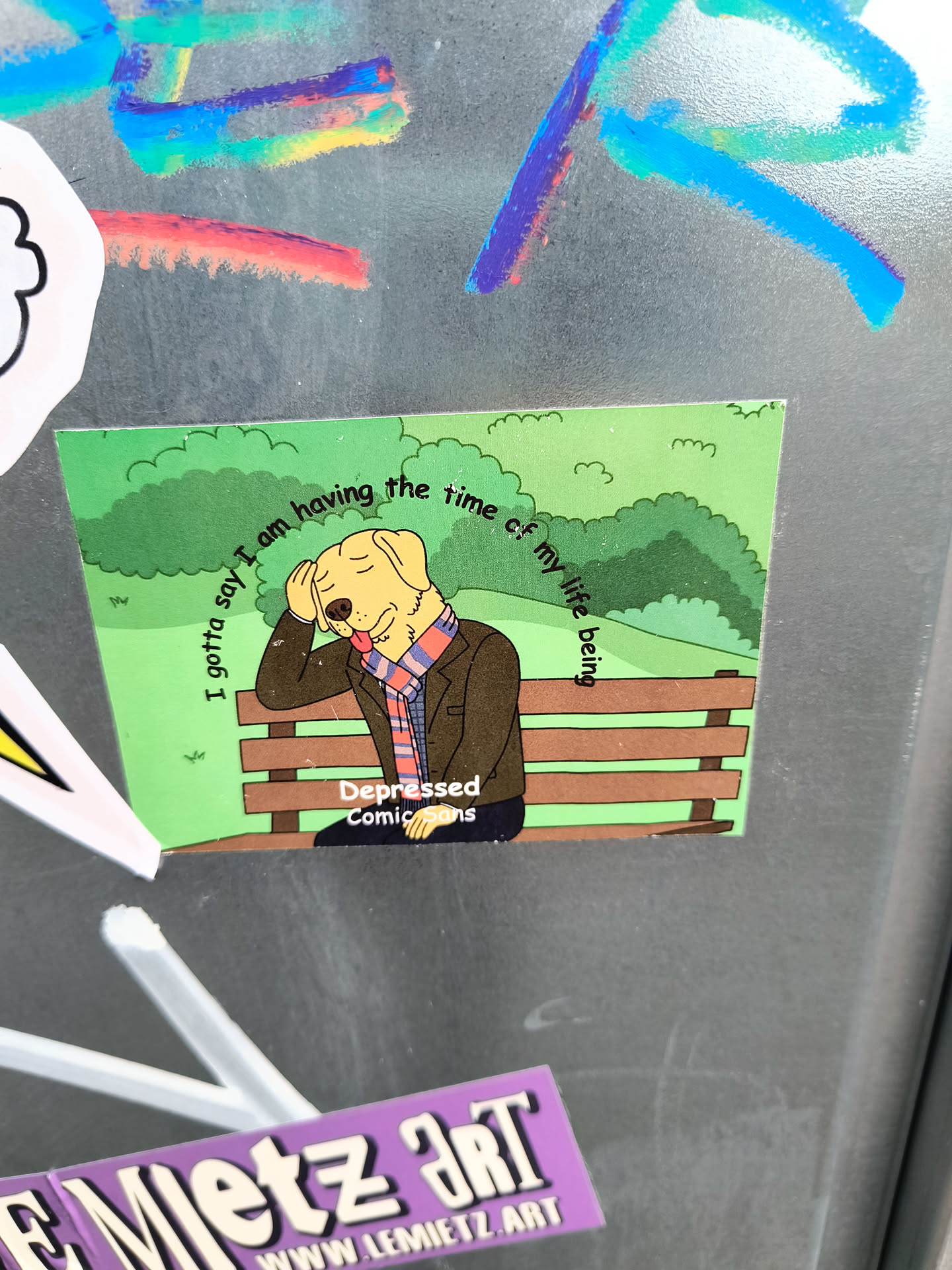 A sticker featuring a cartoon dog sitting on a park bench with text that says, "I gotta say I am having the time of my life being depressed." The sticker has a green background and is created by Depressed Comic Sans. There is also another sticker from www.lemietz.art visible in the photo.