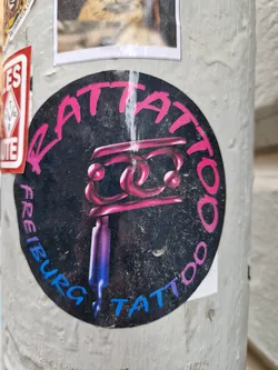 Round sticker with a tattoo machine and the text "Rat Tattoo Freiburg Tattoo" in pink and blue. The sticker has a grunge aesthetic.