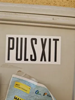 A minimalistic sticker with the text "PULS XIT" in bold, uppercase, sans-serif font. The sticker is white with black text and is affixed to a light beige surface.