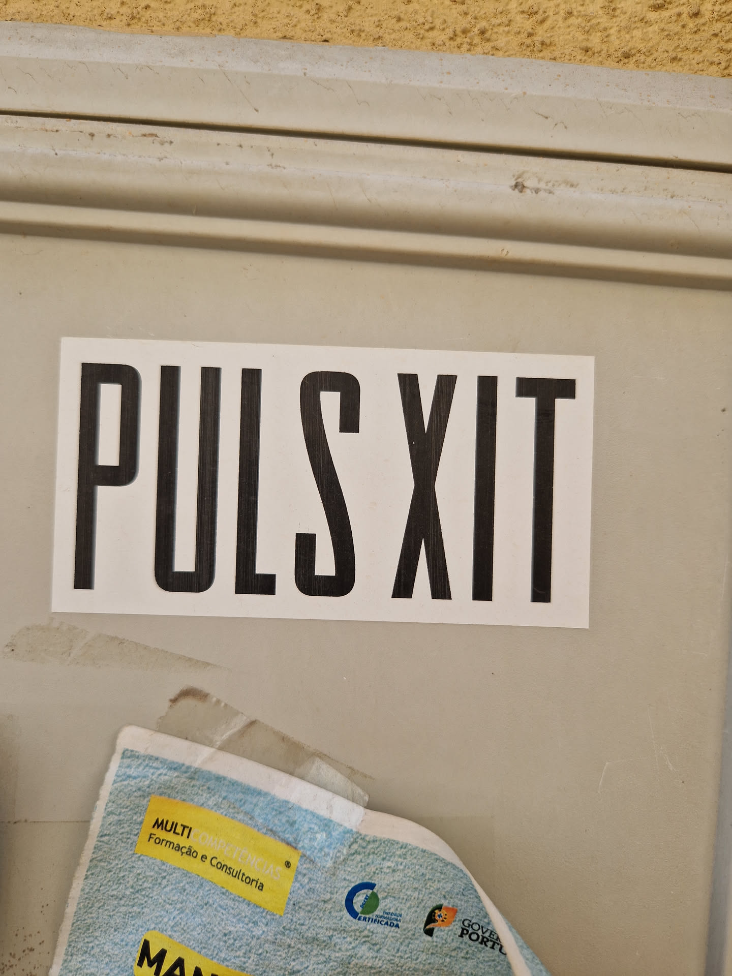 A minimalistic sticker with the text "PULS XIT" in bold, uppercase, sans-serif font. The sticker is white with black text and is affixed to a light beige surface.