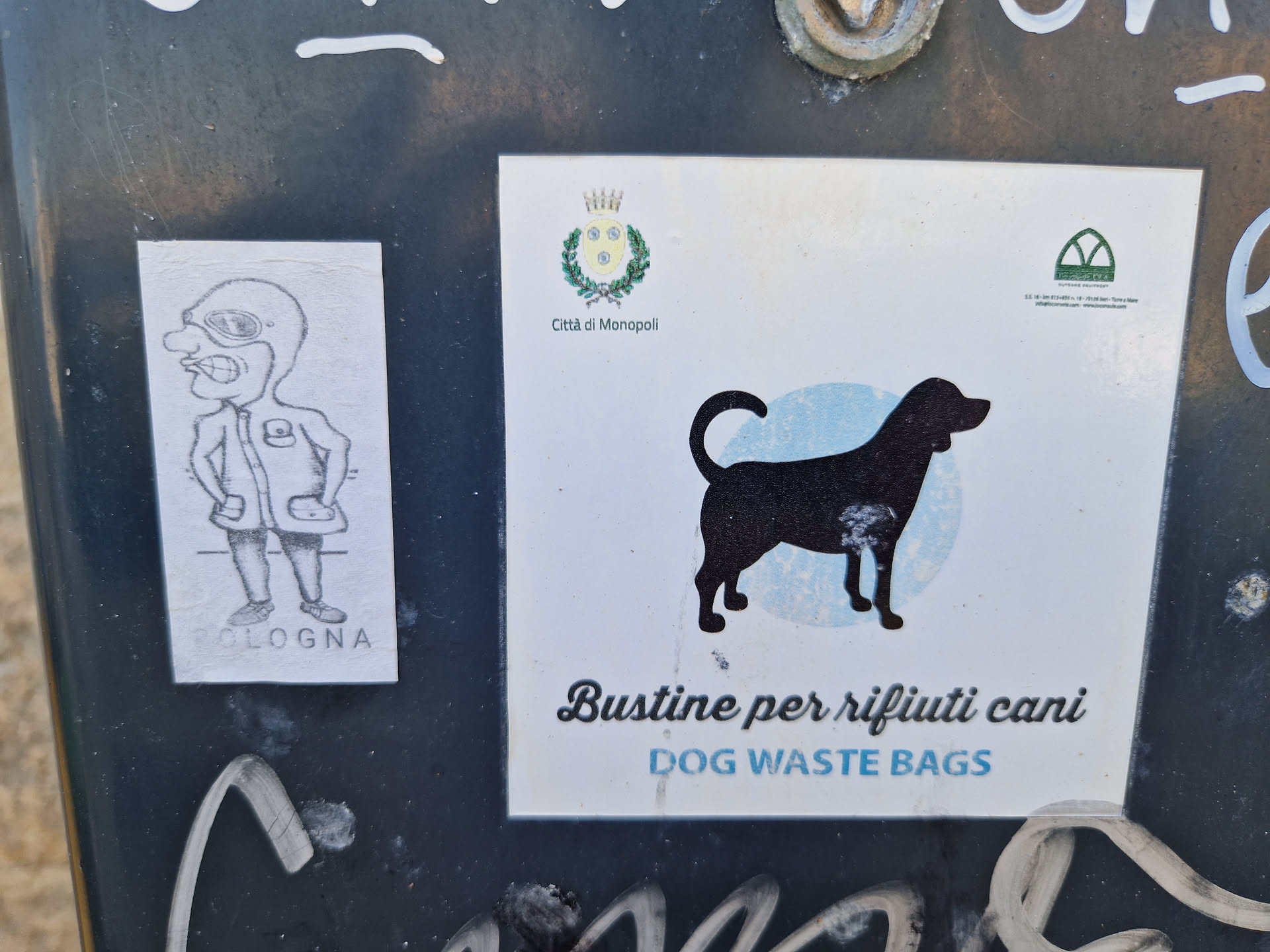 A sticker indicating the availability of dog waste bags.  The sticker features a silhouette of a dog and the text "Bustine per rifiuti cani" (Dog waste bags) in Italian and English.  A smaller sticker to its left shows a cartoon drawing of a man.