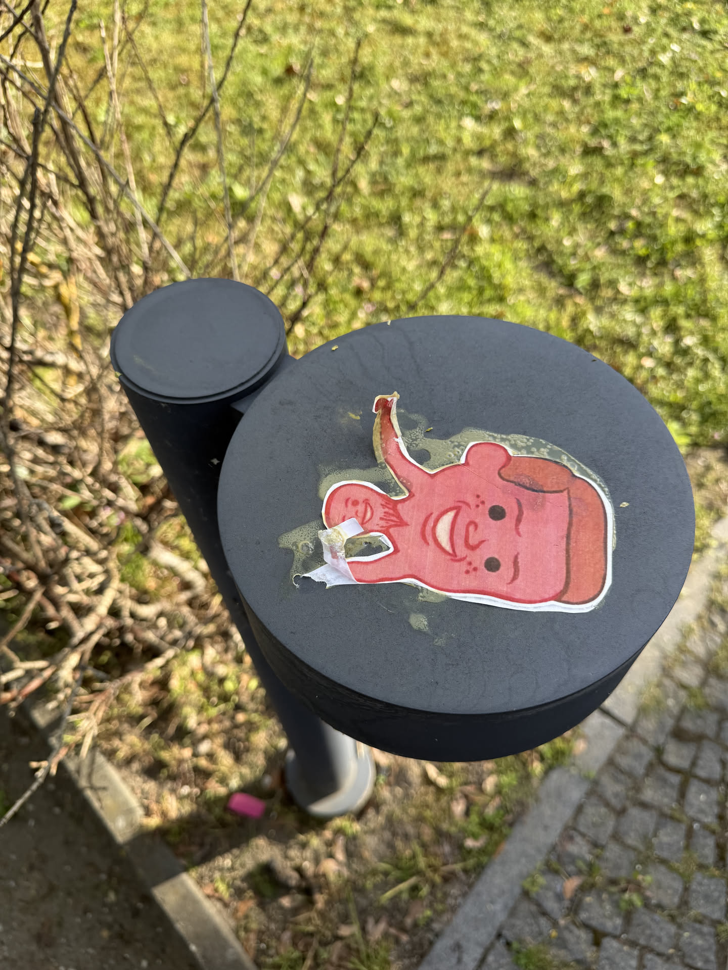 The sticker features a stylized, cartoonish red character, possibly a dog or an abstract creature, with a large, open-mouthed smile revealing white teeth. It has two simple black dot eyes and several small black dots resembling freckles on its face. The character's head is predominantly red with a white outline, and there's some brown shading around its snout and ear. One ear or part of its head extends upwards in an elongated, curved shape. The sticker shows signs of wear, with some peeling and visible yellowish-green adhesive residue around its edges, indicating it has been exposed to the elements.
