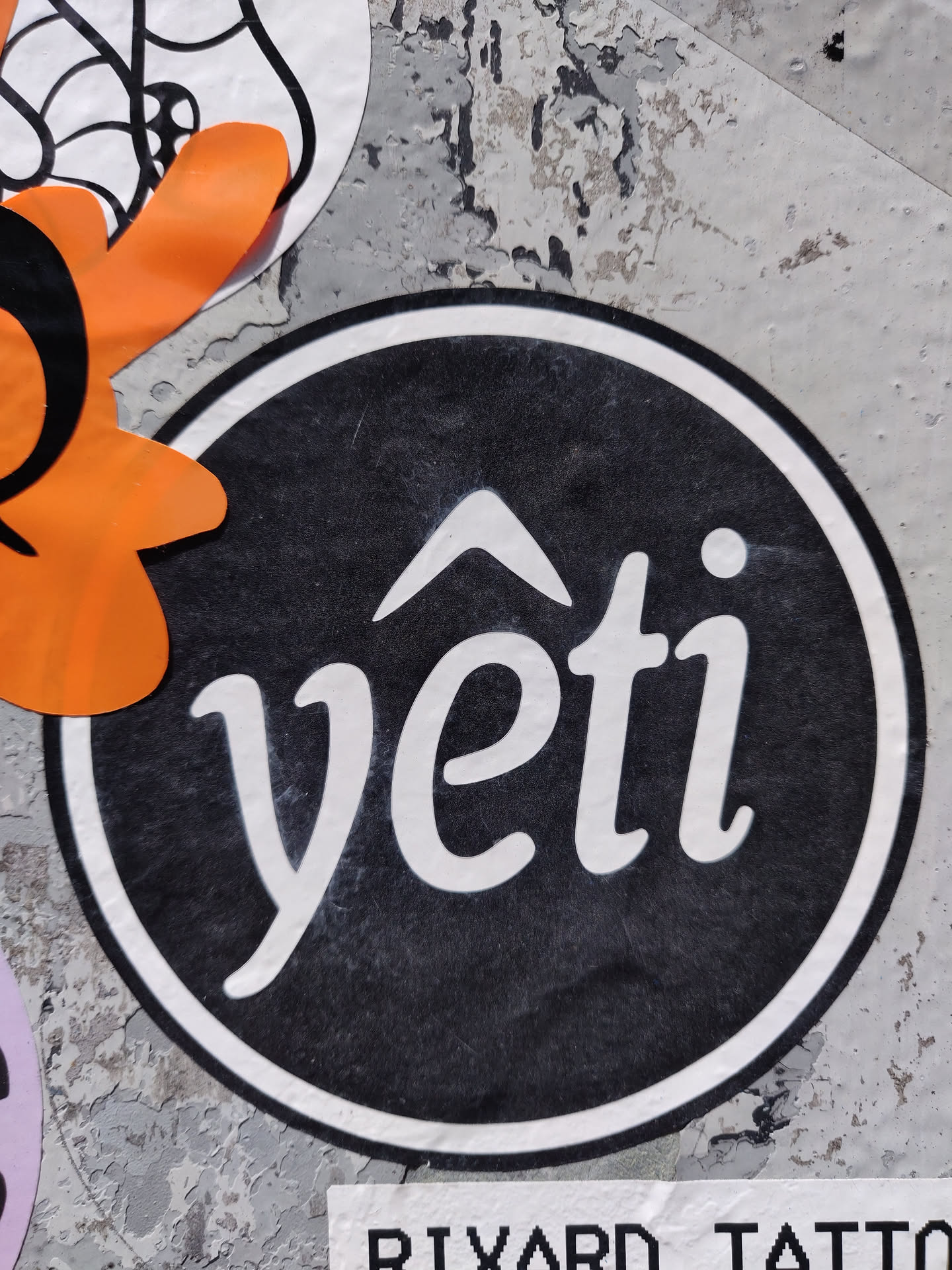 A round black and white sticker with the word "yeti" written in a stylized script font. There's a small upward-pointing arrow above the word. The sticker is affixed to a weathered gray surface.