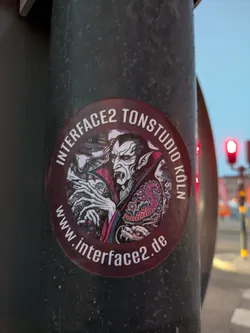 The sticker is circular with a dark burgundy border. It features a detailed illustration of a vampire with sharp fangs, pale skin, and wild white hair. The vampire is depicted with its mouth open, revealing fangs, and has prominent pointed ears. It has intricate tattoos on its chest, including a colorful serpentine design and floral elements. The vampire's hands are visible, with long, slender fingers. Behind the vampire, there are hints of a bat and possibly a moonlit sky. The overall color scheme is dominated by shades of grey, white, black, red, and pink, giving it a gothic and somewhat menacing aesthetic.