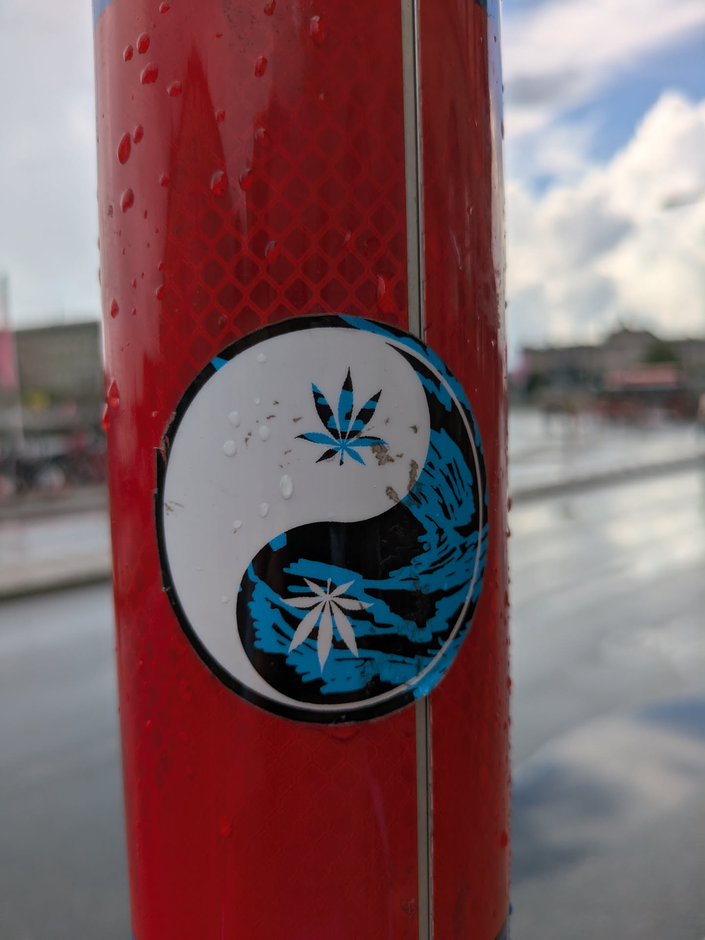 The sticker is circular, displaying a stylized yin-yang symbol. The upper, white half of the yin-yang contains a blue cannabis leaf. The lower, black half features a white cannabis leaf and is filled with an abstract, wave-like or brushstroke pattern in a bright, electric blue. The sticker is affixed to a red, reflective pole and is covered in small water droplets, indicating it's wet. There are also some visible scuffs and dirt marks, particularly on the white section.