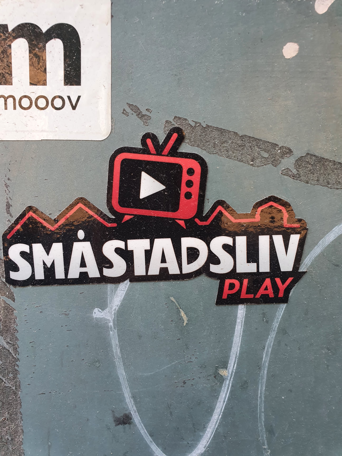 A sticker featuring a retro TV with a play button and the text 'Småstadsliv PLAY'. The design is stylized with a cityscape silhouette behind the TV.  The colors are primarily black, red, and gold.