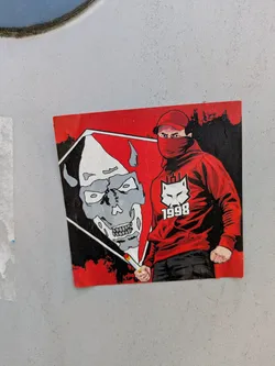 The sticker is red and black. It depicts a person with a red mask and cap, wearing a red hoodie with a white logo and the year "1998". Next to the person is a stylized skull.