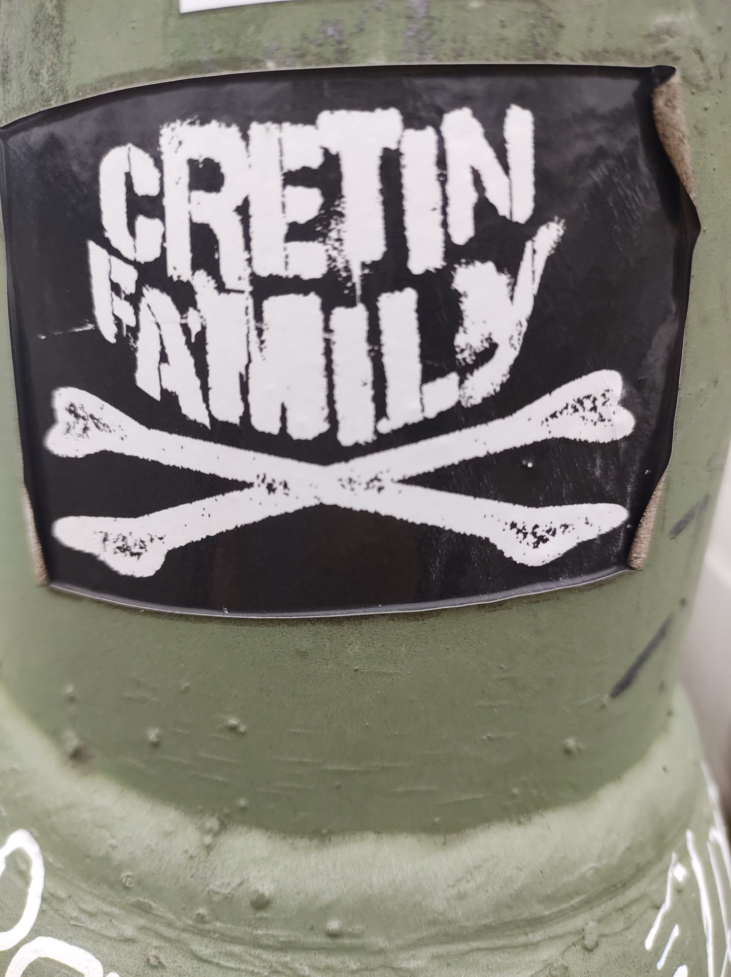 Black and white sticker with the text "CRETIN FAMILY" in a stylized font, along with a graphic of crossed bones. The sticker is affixed to a green surface.