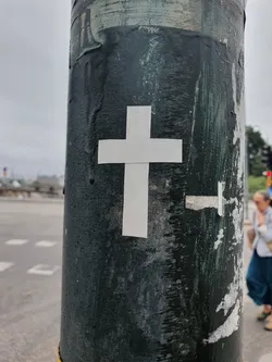 A minimalist sticker featuring a white cross is affixed to a dark green pole. The pole shows signs of wear and tear, with remnants of other stickers visible.