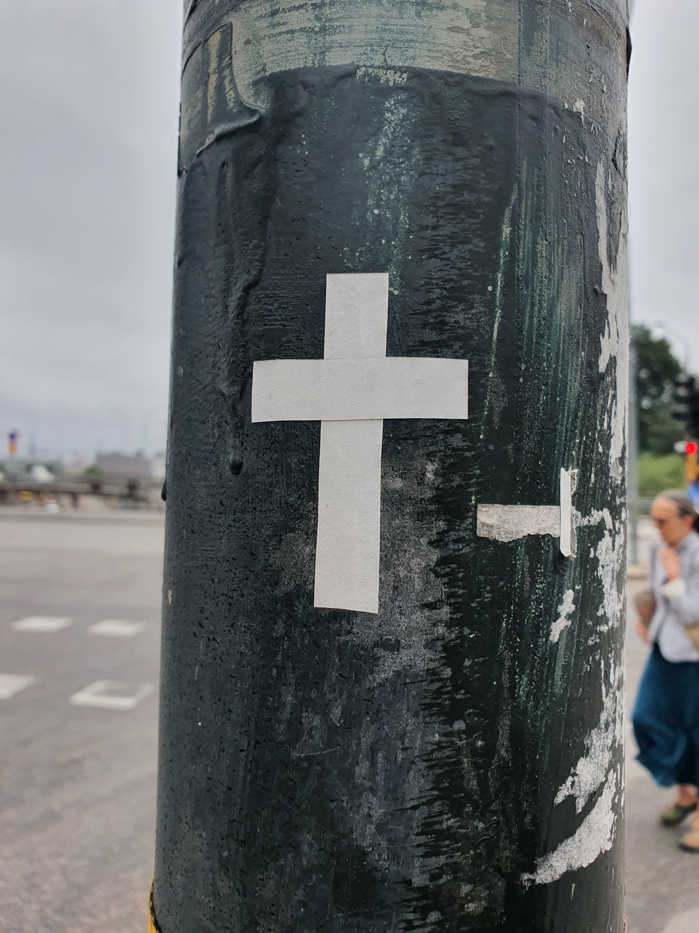 A minimalist sticker featuring a white cross is affixed to a dark green pole. The pole shows signs of wear and tear, with remnants of other stickers visible.