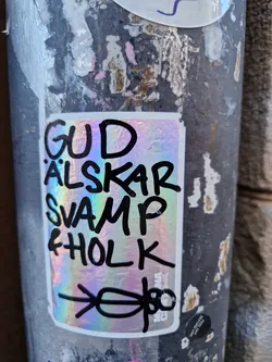 A holographic sticker with the text "Gud älskar svamp & holk" (God loves mushrooms and fungi) and a drawing of two figures at the bottom. The sticker is attached to a pole.