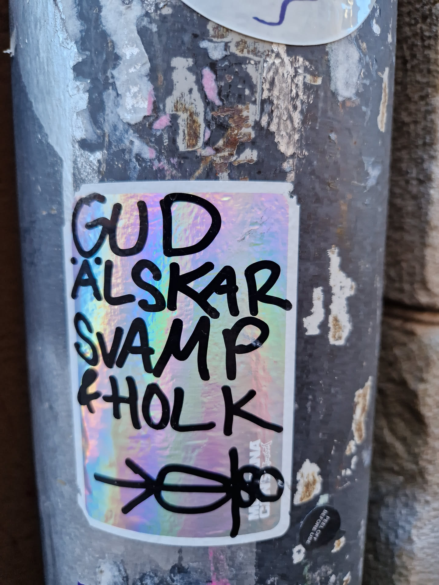 A holographic sticker with the text "Gud älskar svamp & holk" (God loves mushrooms and fungi) and a drawing of two figures at the bottom. The sticker is attached to a pole.
