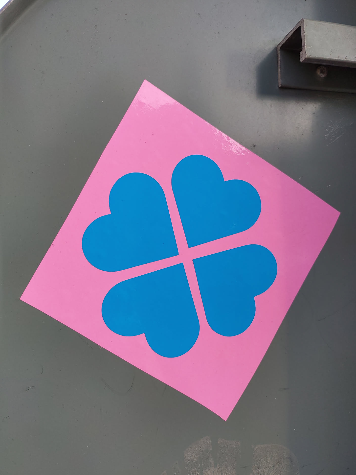 A diamond-shaped sticker with a pink background and a blue four-leaf clover design made of four hearts. The sticker is affixed to a gray surface.