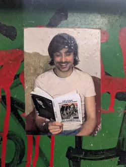 Street sticker The sticker features a photograph of a smiling person with dark, wavy hair, wearing a light-colored t-shirt and blue jeans. The person is holding open a book. The book's cover shows a black and white image of a woman and text that reads 'HELENA HOOGENKAMP HET AANBIDDEN VAN LOUIS CLAUS'. The sticker is surrounded by a green background with abstract splatters and drips of red and black paint, giving it a street art or graffiti aesthetic.