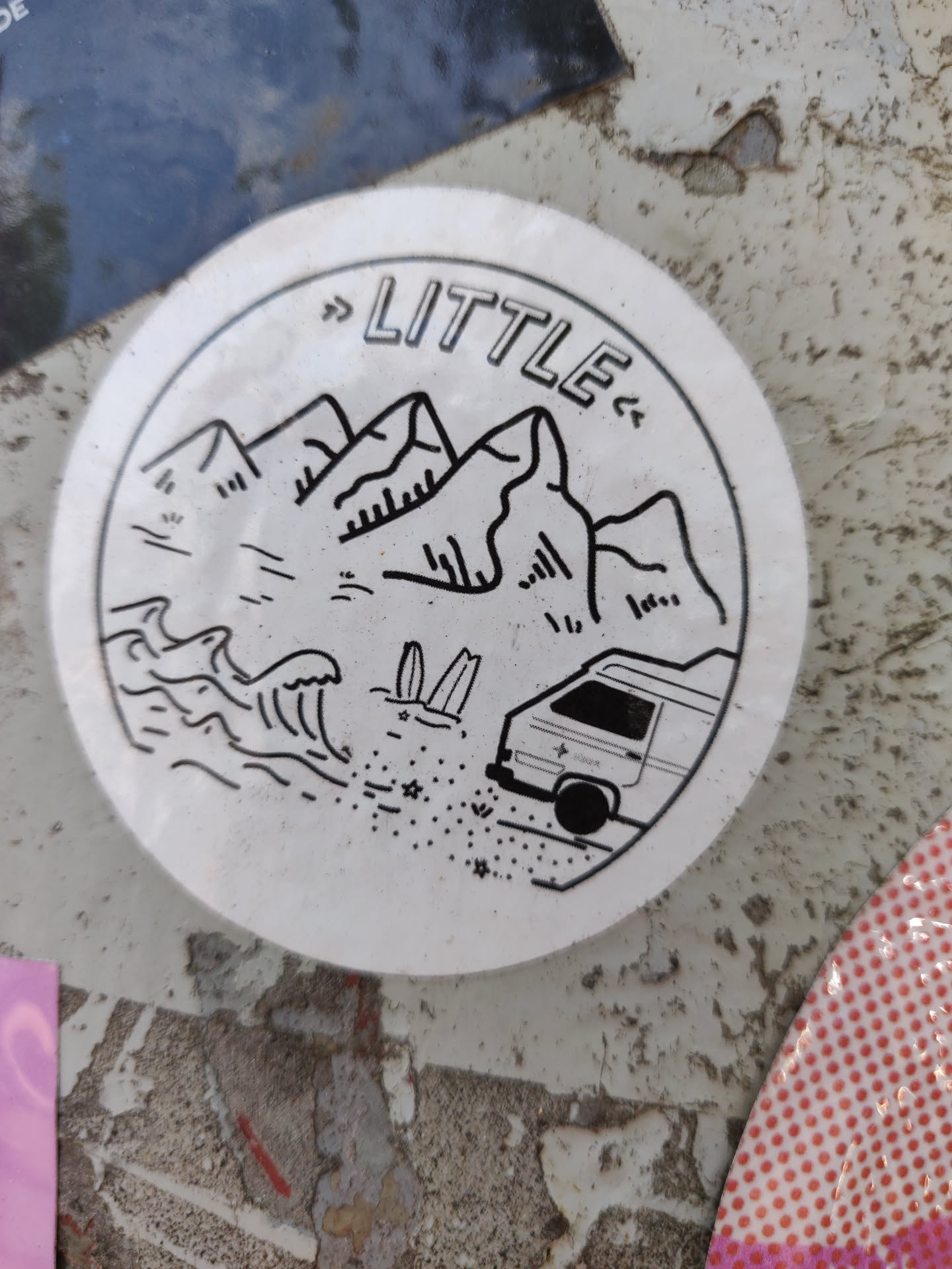 Round sticker with a minimalist design featuring mountains, ocean waves, surfboards, and a van. The word "LITTLE" is written in the center.