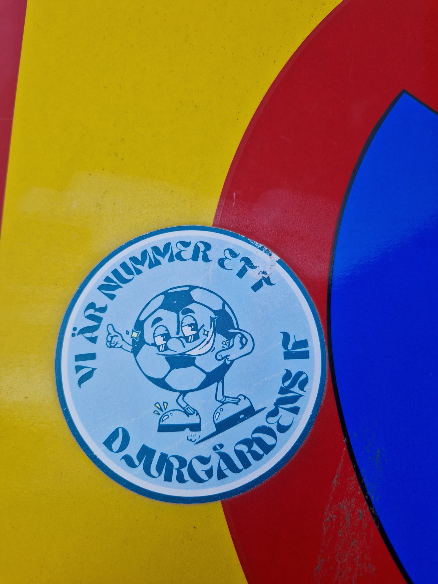 A round sticker featuring a cartoon soccer ball character with the text "Vi är Nummer Ett Djurgårdens IF" which translates to "We are number one Djurgårdens IF".  The sticker is affixed to a surface with yellow, red, and blue sections.