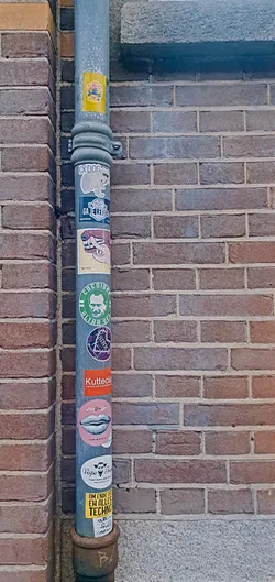 A collection of various stickers is adhered to a metal pipe set against a brick wall. The stickers vary in design and color, ranging from text-based messages to graphic images. Some of the stickers appear to be related to music or events, while others are more abstract or humorous in nature.