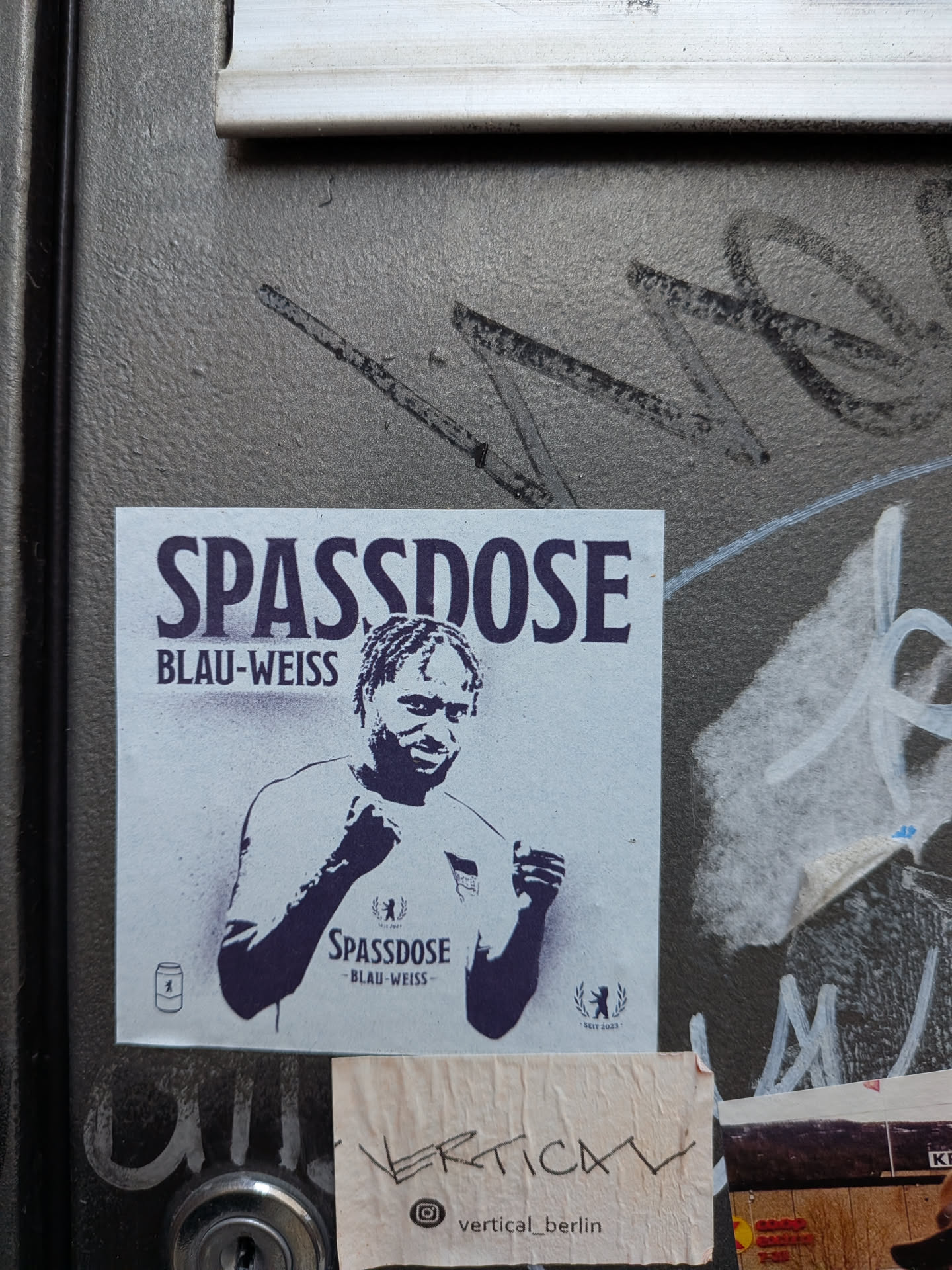 A square paper sticker printed in dark navy blue or purple monochrome on an off-white background. At the top, bold capital letters read 'SPASSDOSE' above 'BLAU-WEISS'. In the center is a high-contrast graphic representation of a Hertha BSC footballer pumping his fists in celebration, wearing a jersey customized with the same 'SPASSDOSE' text. In the lower left corner is a small icon of a beverage can with the Berlin bear emblem, while the lower right corner features the Berlin bear enclosed in a laurel wreath with 'SEIT 2023'.