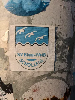 A rectangular sticker with a rounded bottom, resembling a crest or shield. The upper half features a light blue background with three white seagulls in flight. Below them, a black wavy line separates this section from the white lower half. The white section contains text in blue: "SV Blau-Weiß" in a straight line and "SCHOLLENE" curved underneath. The sticker is bordered by a thin black outline and is adhered to a textured, weathered pole or wall, surrounded by remnants of other old stickers.