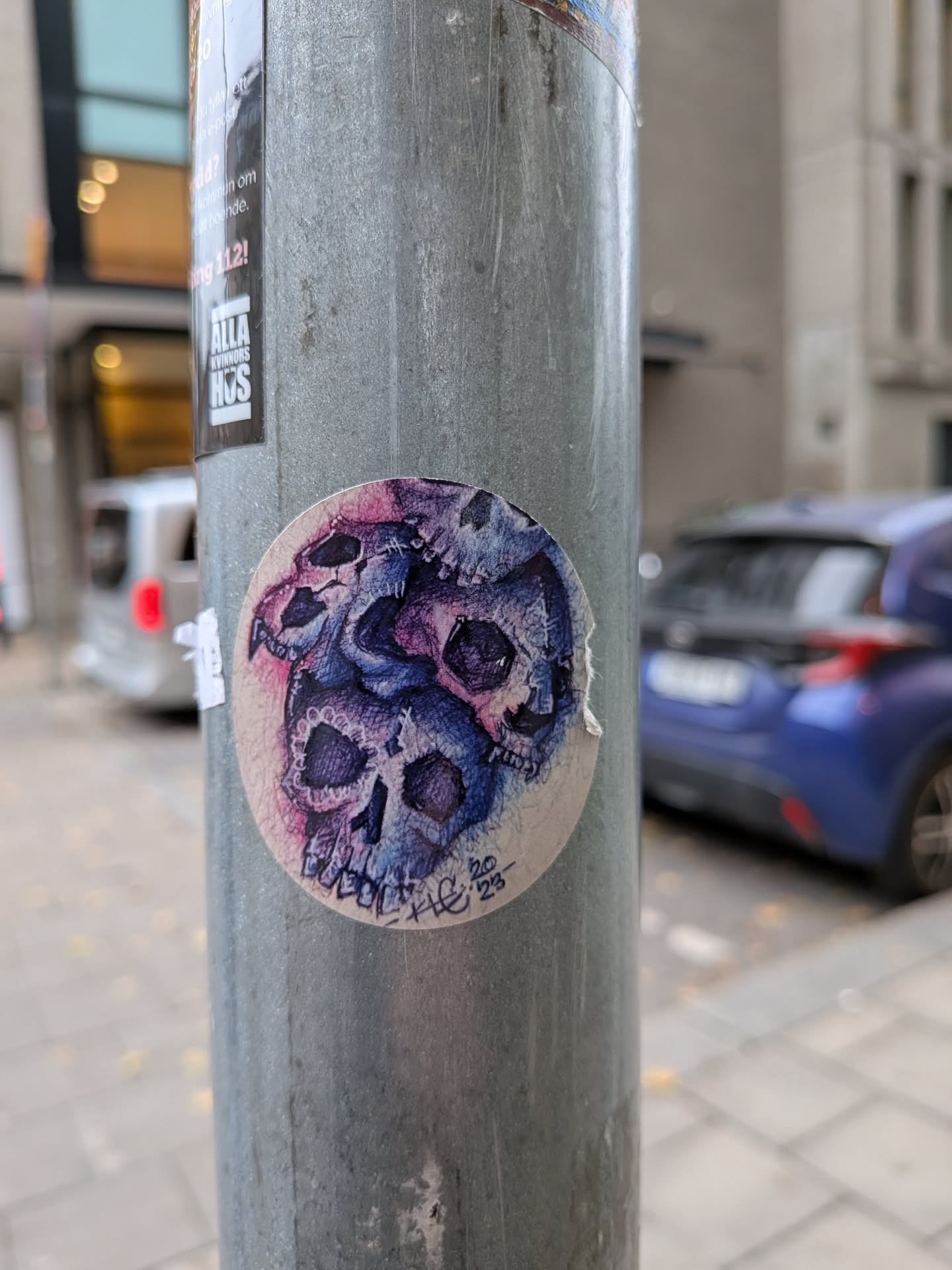 The sticker is circular and features a detailed illustration of multiple skulls intertwined. The skulls are depicted in shades of deep blue, purple, and pink, with intricate details in their eye sockets and teeth. The overall style is somewhat gothic and macabre. There is some handwritten text and numbers in the lower right corner of the sticker.