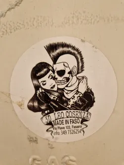 Round sticker featuring a monochrome illustration of a skull with a mohawk embracing a girl with a bob haircut.  The sticker has the artist's name and contact information. 