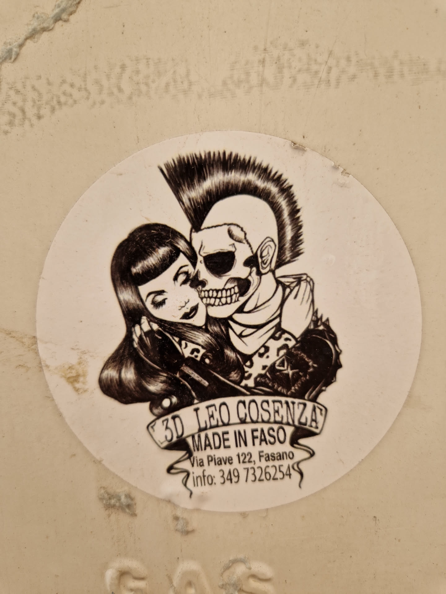 Round sticker featuring a monochrome illustration of a skull with a mohawk embracing a girl with a bob haircut.  The sticker has the artist's name and contact information. 