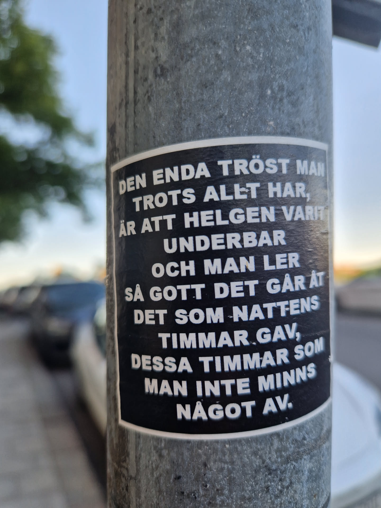This sticker features a passage of text, apparently in Swedish, and it seems to reflect some amount of light, giving it a metallic sheen. The sticker is stuck to a grey metal pole. The text on the sticker reads: "DEN ENDA TRÖST MAN TROTS ALLT HAR, ÄR ATT HELGEN VARIT UNDERBAR OCH MAN LER SÅ GOTT DET GÅR ÅT DET SOM NATTENS TIMMAR GAV, DESSA TIMMAR SOM MAN INTE MINNS NÅGOT AV."