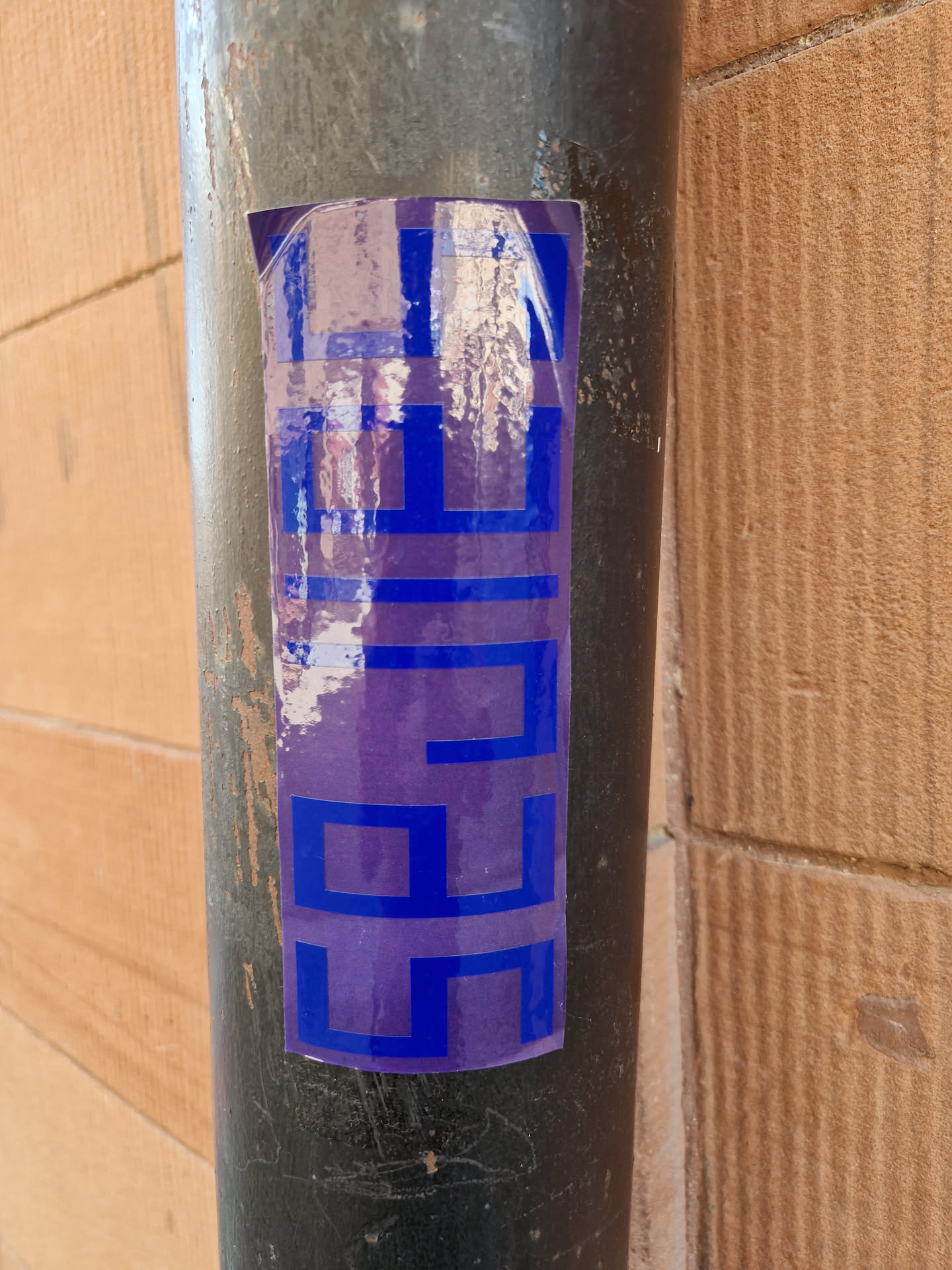 A sticker with a repeating geometric pattern in shades of purple and blue is affixed to a dark-colored pipe. The sticker appears to be made of a slightly transparent material, allowing some of the pipe's surface to show through. The background is a light brown brick wall.