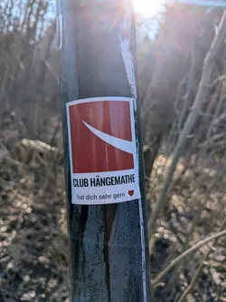 The sticker is rectangular with a white border. Its main design features a large red background with a prominent, thick white diagonal stripe or 'swoosh' running from the top-left towards the bottom-right. Below this graphic, in bold black uppercase letters, is the text 'CLUB HÄNGEMATHE'. Beneath that, in smaller black letters, it reads 'hat dich sehr gern', followed by a small red heart icon.