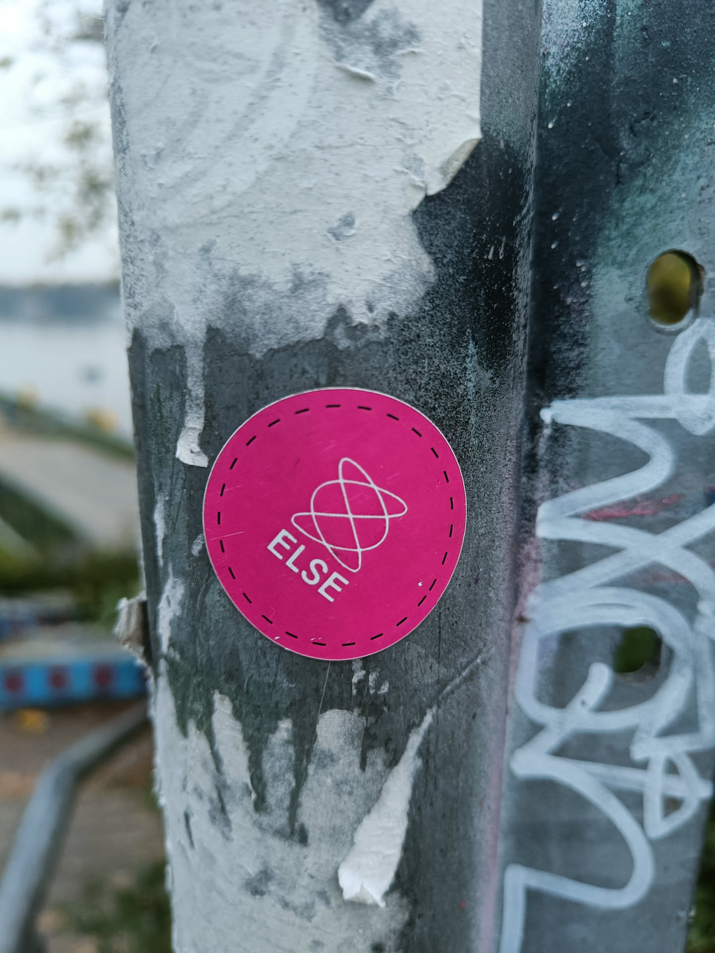 A round pink sticker with the word 'ELSE' in white text and a simple graphic above it.  The sticker has a dashed-line border and is affixed to a weathered metal pole. The background shows remnants of other stickers and graffiti.