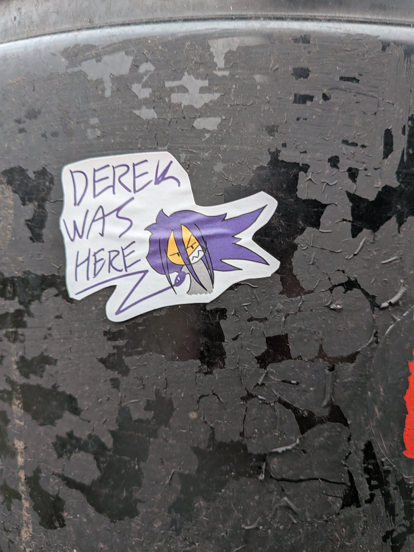 The sticker features a stylized cartoon character with vibrant purple, spiky hair that extends sharply to the right. The character has yellow, almond-shaped eyes with black pupils and an angry, determined expression, showing sharp white teeth. A grey, almost metallic-looking element is visible below the character's face, possibly part of a collar or clothing. To the left of the character, in a handwritten, somewhat jagged purple font, are the words 'DEREK WAS HERE'. The entire design is outlined with a clean white border, giving it a distinct cut-out appearance against the dark, peeling background surface.