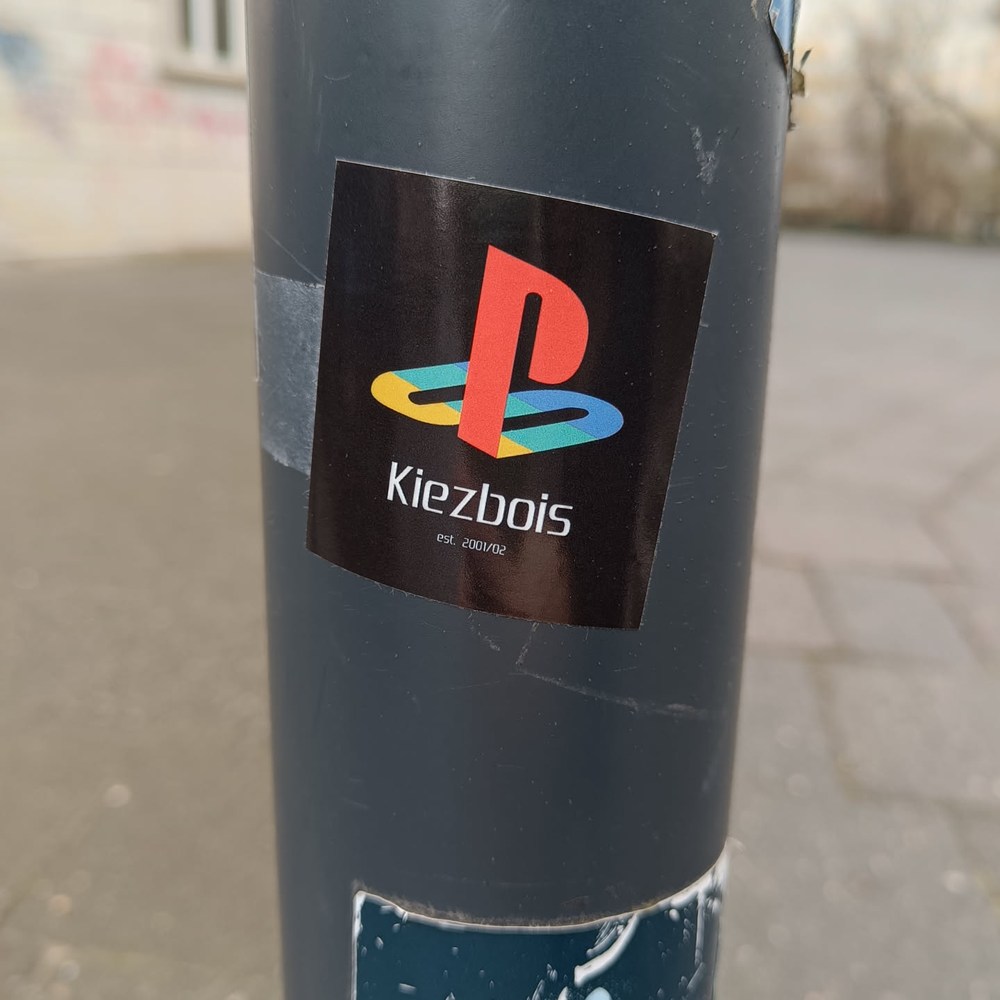 Black square sticker with a stylized Playstation logo and the text 'Kiezbois' underneath.  It also has 'est. 2001/02' in smaller text below the name. The Playstation logo is in red, yellow, blue and green. The sticker appears to be on a gray pole.