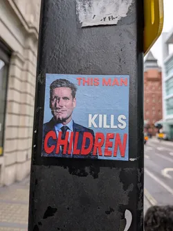 A rectangular sticker adhered to a black pole, featuring a portrait of a man in a suit and tie against a light blue background. The man's face has a vertical, distressed or 'scratched' effect. Bold text is overlaid on the image: 'THIS MAN' in red at the top, 'KILLS' in white below it, and 'CHILDREN' in large red letters at the bottom. The sticker shows some signs of wear and tear, particularly around the edges.