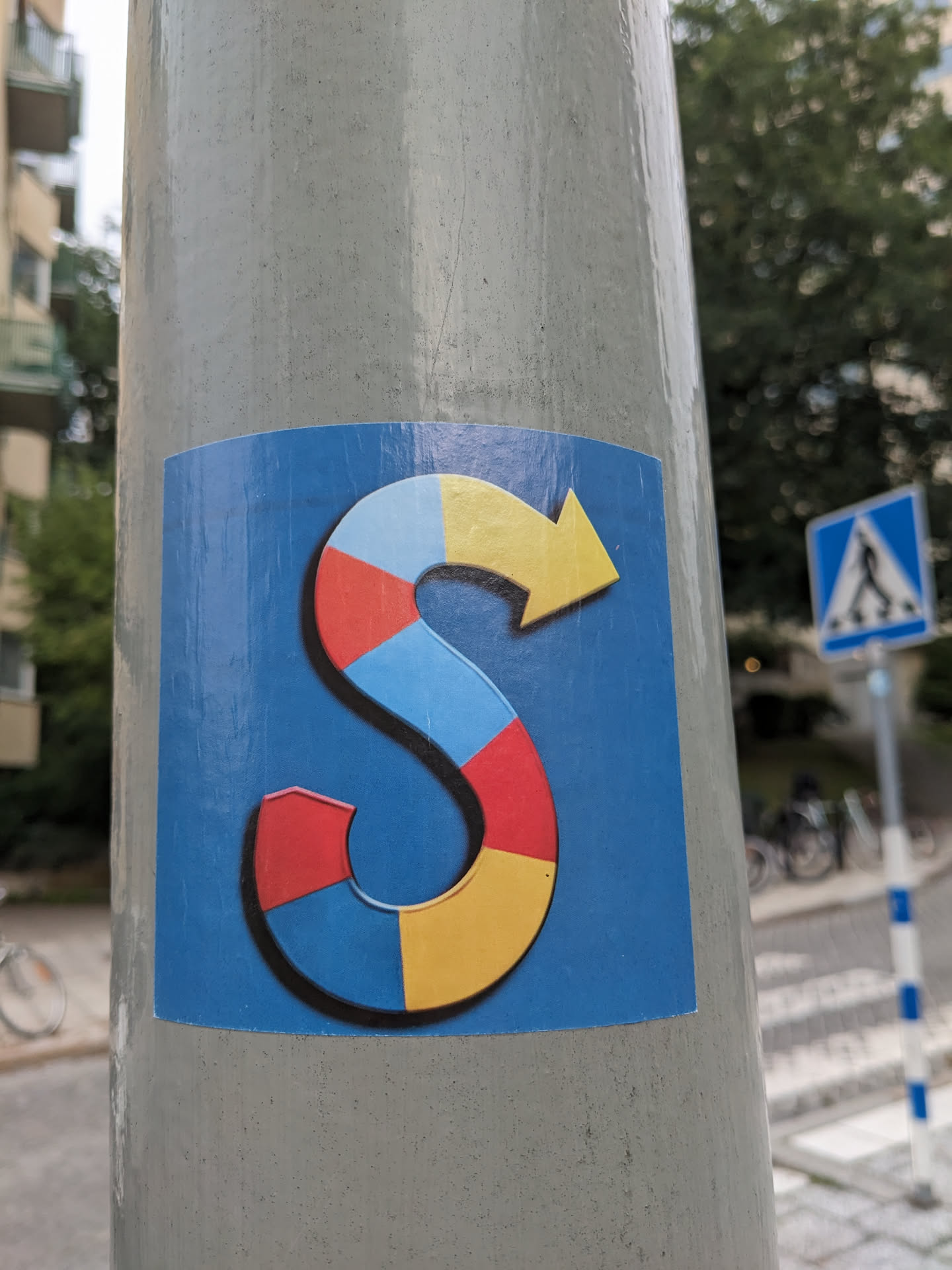 A sticker featuring a stylized letter S formed by segments of red, yellow, blue and light blue. The S has a yellow arrow at its end. The background is blue.