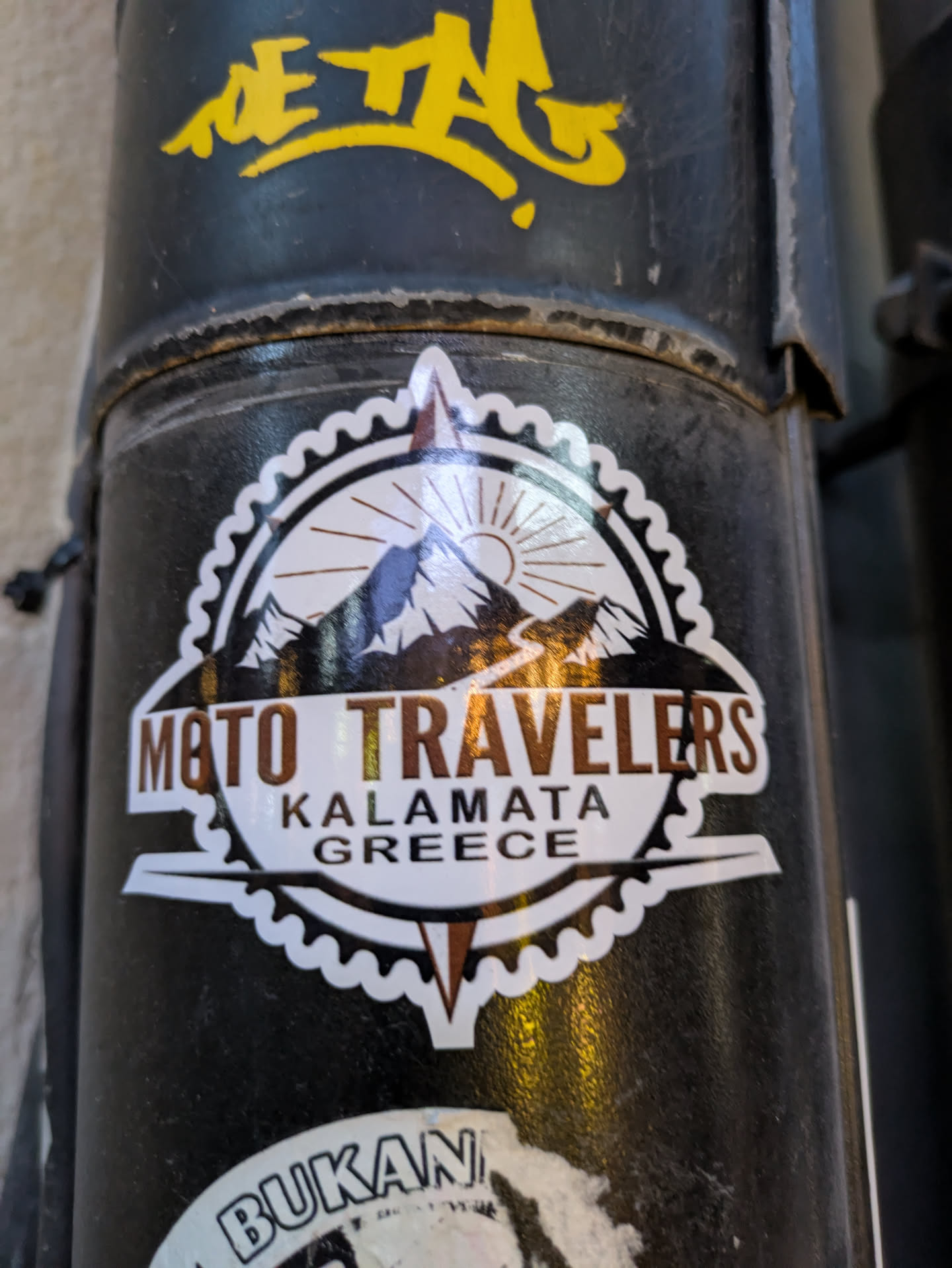 The sticker is circular with a jagged, gear-like outer edge in white. The central design features a stylized mountain range under a sun with rays, depicted in shades of brown and white. Below the mountains, the text "MOTO TRAVELERS" is prominently displayed in a metallic brown font with a white outline. Underneath this, in smaller white text, are the words "KALAMATA GREECE". The overall aesthetic suggests a travel or adventure theme, possibly related to motorcycles.