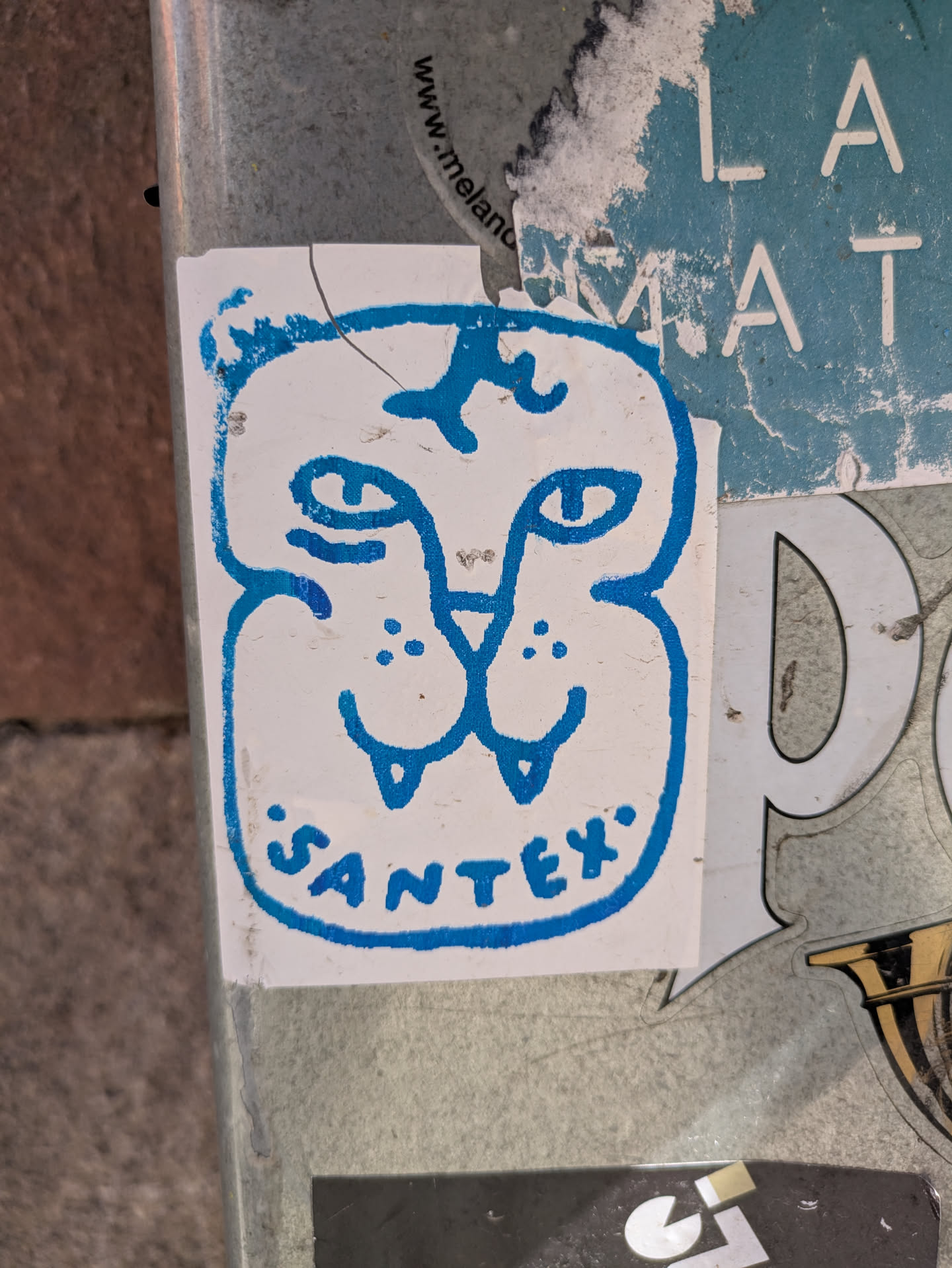 A small, white, square sticker features a blue line drawing of a stylized cat face. The drawing has simple features: eyes, nose, whiskers, and a star above its head. Below the face, the word "Santex." is written in a rounded sans-serif font, also in blue. The sticker appears to be on a pole or wall outdoors.