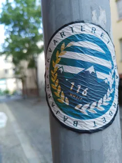A round sticker featuring the text "Kvarteret 116 MMXIX" encircled in a laurel wreath design. The main color scheme is teal and white. The sticker appears to be affixed to a grey pole outdoors.