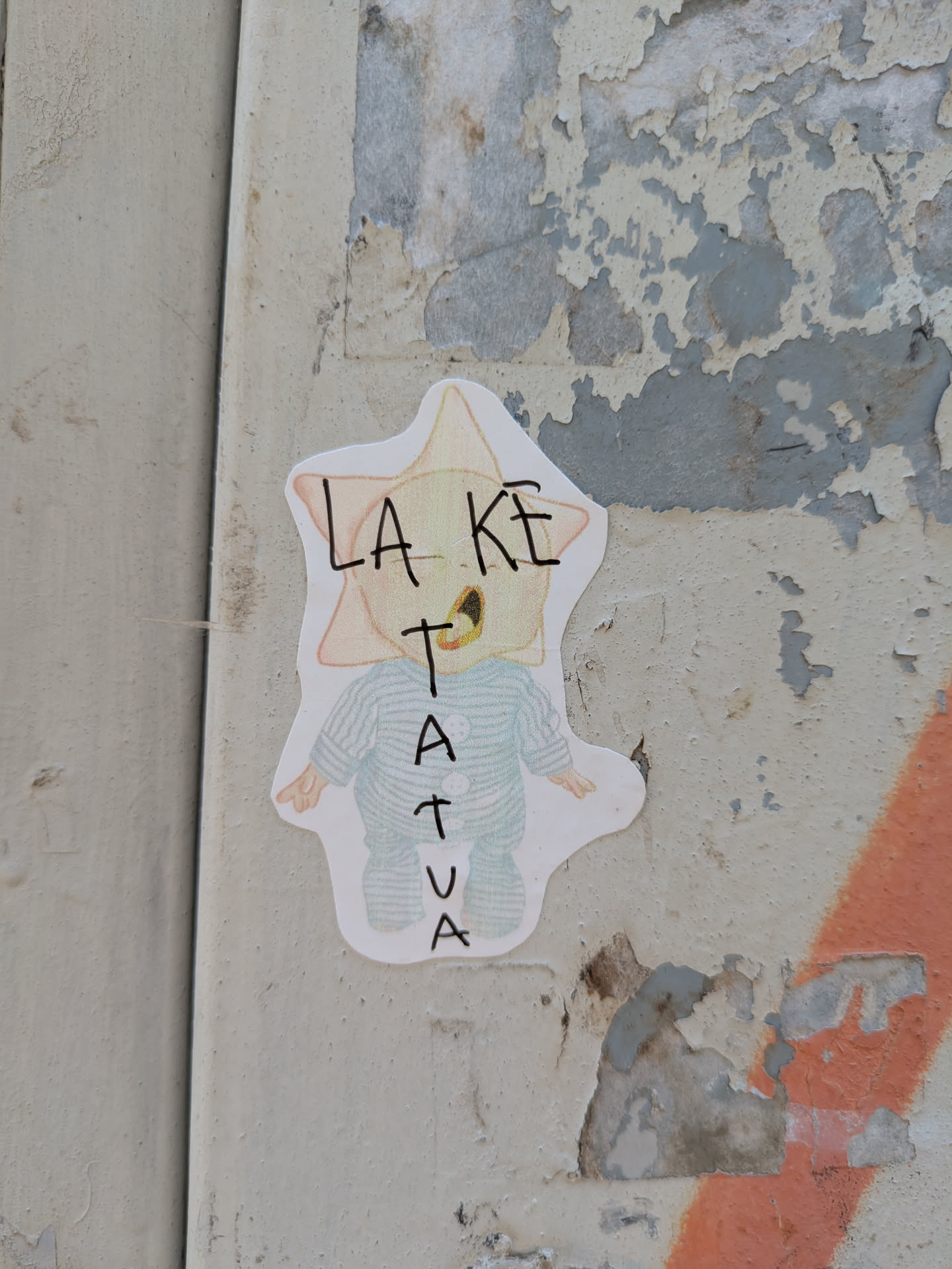 This sticker showcases a cartoonish figure with a star-shaped head. The character wears striped pajamas and has the word "LAKETATUA" scrawled on its face. It's pasted on a weathered surface with peeling paint.
