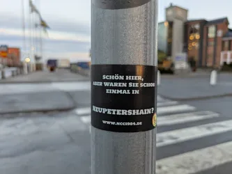 A black sticker on a metal pole with text in German.  The text translates to: "Nice here, but have you ever been to Neupetershain?"  A website address is also included.