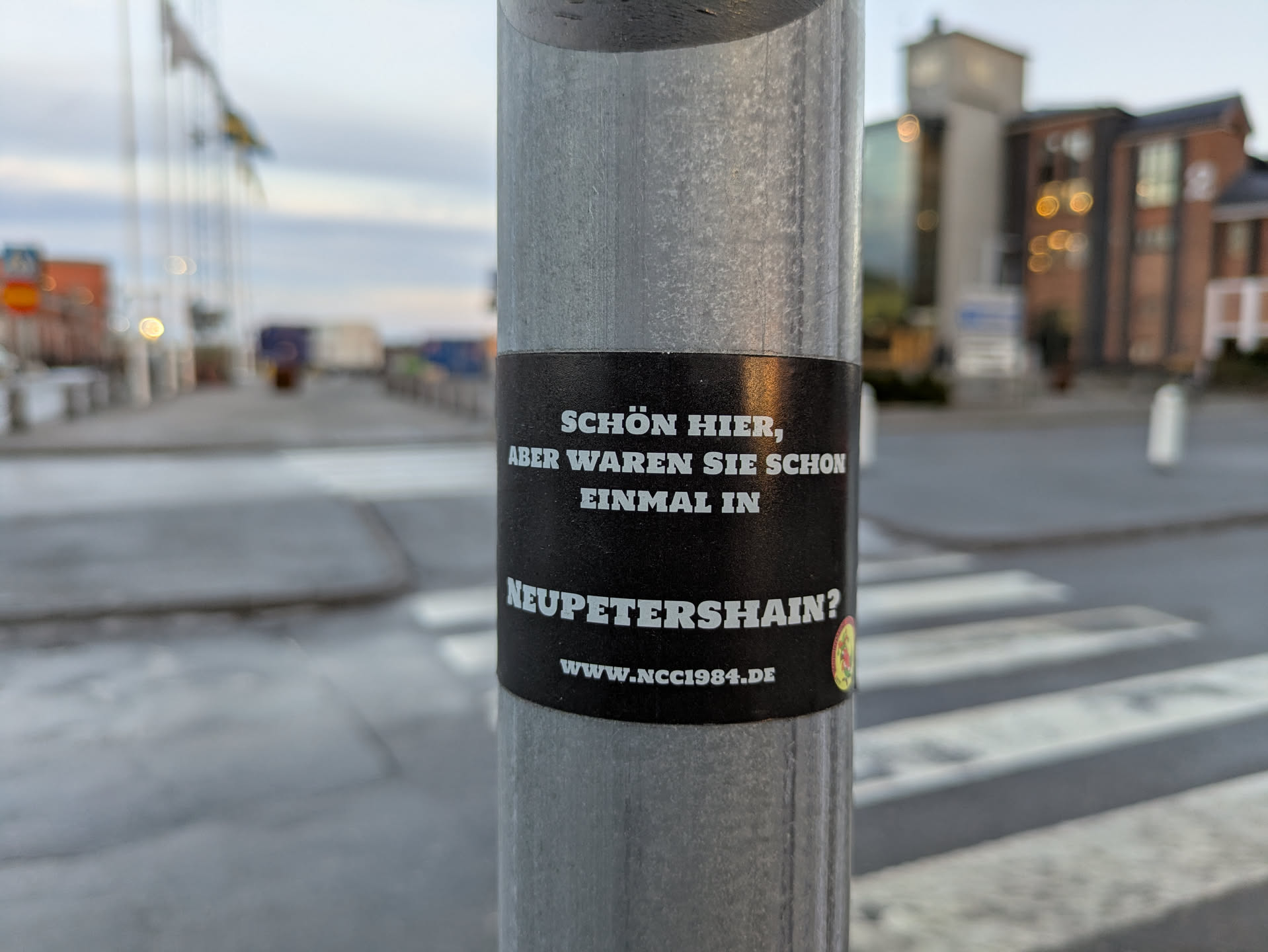 A black sticker on a metal pole with text in German.  The text translates to: "Nice here, but have you ever been to Neupetershain?"  A website address is also included.
