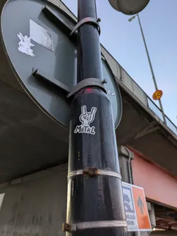 A sticker featuring a hand making the metal horns sign with the word "METAL" underneath it, affixed to a pole under a bridge.