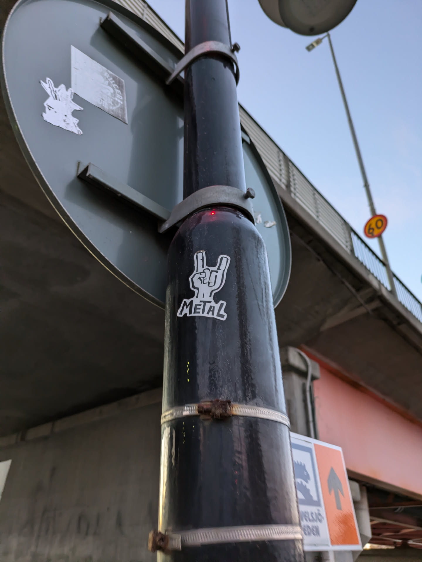 A sticker featuring a hand making the metal horns sign with the word "METAL" underneath it, affixed to a pole under a bridge.
