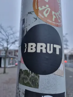 A black circular sticker with the word "BRUT" in a bold, sans-serif, uppercase, off-white font. The sticker is affixed to a pole covered in other stickers.