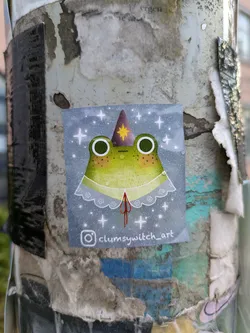 A square sticker prominently features a light green frog with large dark eyes, adorned with a purple wizard hat with a yellow star and a delicate white lace collar. The background is a soft blue-grey, sprinkled with numerous white stars and small white dots, creating a celestial or magical ambiance. The sticker is affixed to a weathered and textured pole, surrounded by remnants of other old papers and adhesives.