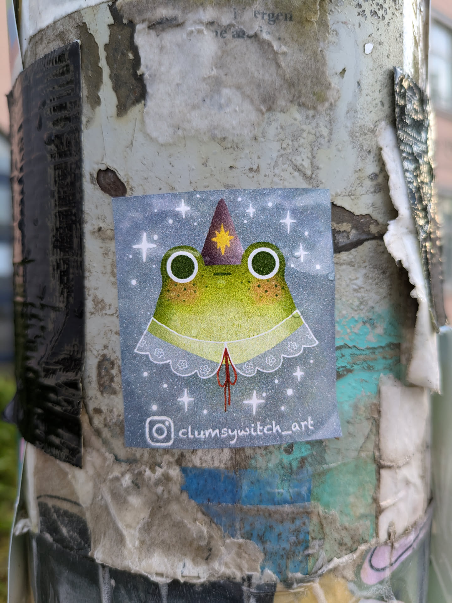 A square sticker prominently features a light green frog with large dark eyes, adorned with a purple wizard hat with a yellow star and a delicate white lace collar. The background is a soft blue-grey, sprinkled with numerous white stars and small white dots, creating a celestial or magical ambiance. The sticker is affixed to a weathered and textured pole, surrounded by remnants of other old papers and adhesives.