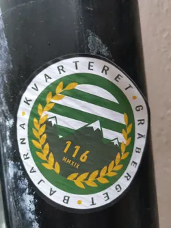 Round green sticker with gold details. It has the text "Kvarteret Gråberget" written around a central design, which shows three mountain peaks, three horizontal stripes and a laurel wreath. At the center, there is the number 116 and the year MMXIX (2019).